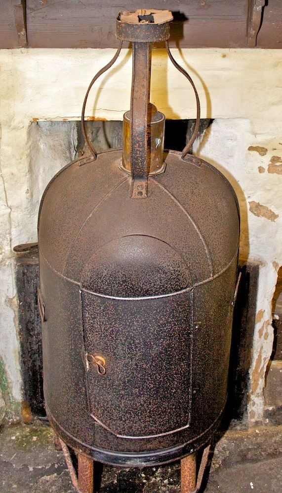 Photo of the Dutch Oven in front of a fireplace in the Cottage Museum. It's a metal structure that stands about waist high on 4 short legs. The body of the oven is semicurcular and open at the back, facing the fire. There is a door in the front. At the top a frame holds a brass cylinder which is a clockwork jack from which meat can be suspended inside the oven.